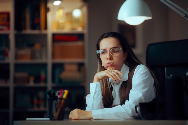 A woman wearing glasses and a white shirt sits at a desk, resting her chin on her hand and looking thoughtful or bored—perhaps pondering government hiring prospects. Shelves with books and a lamp are visible in the background.