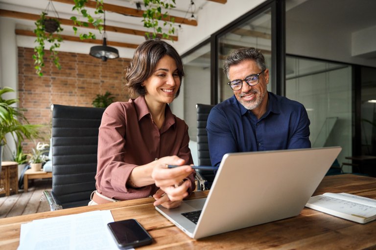 Two smiling colleagues sit at a desk in a modern office, looking at a laptop together. One person points at the screen, reflecting their collaborative culture. Papers, a phone, and plants decorate the welcoming workspace.