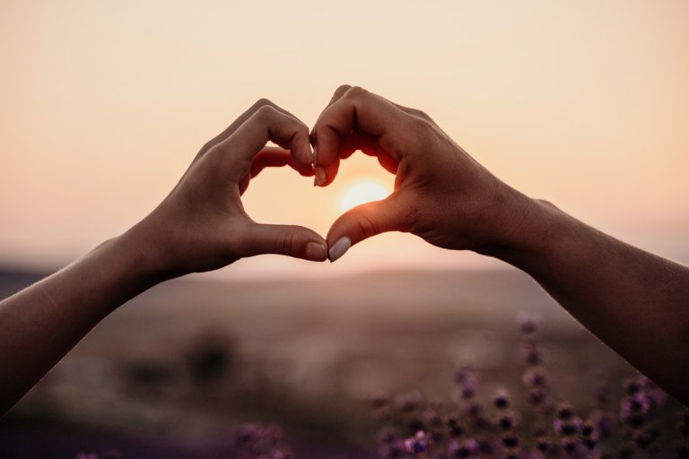 Two hands form the shape of a heart against a sunset, with the sun centered inside the heart shape—capturing a moment filled with faith and warmth, set against a blurred landscape in the background.