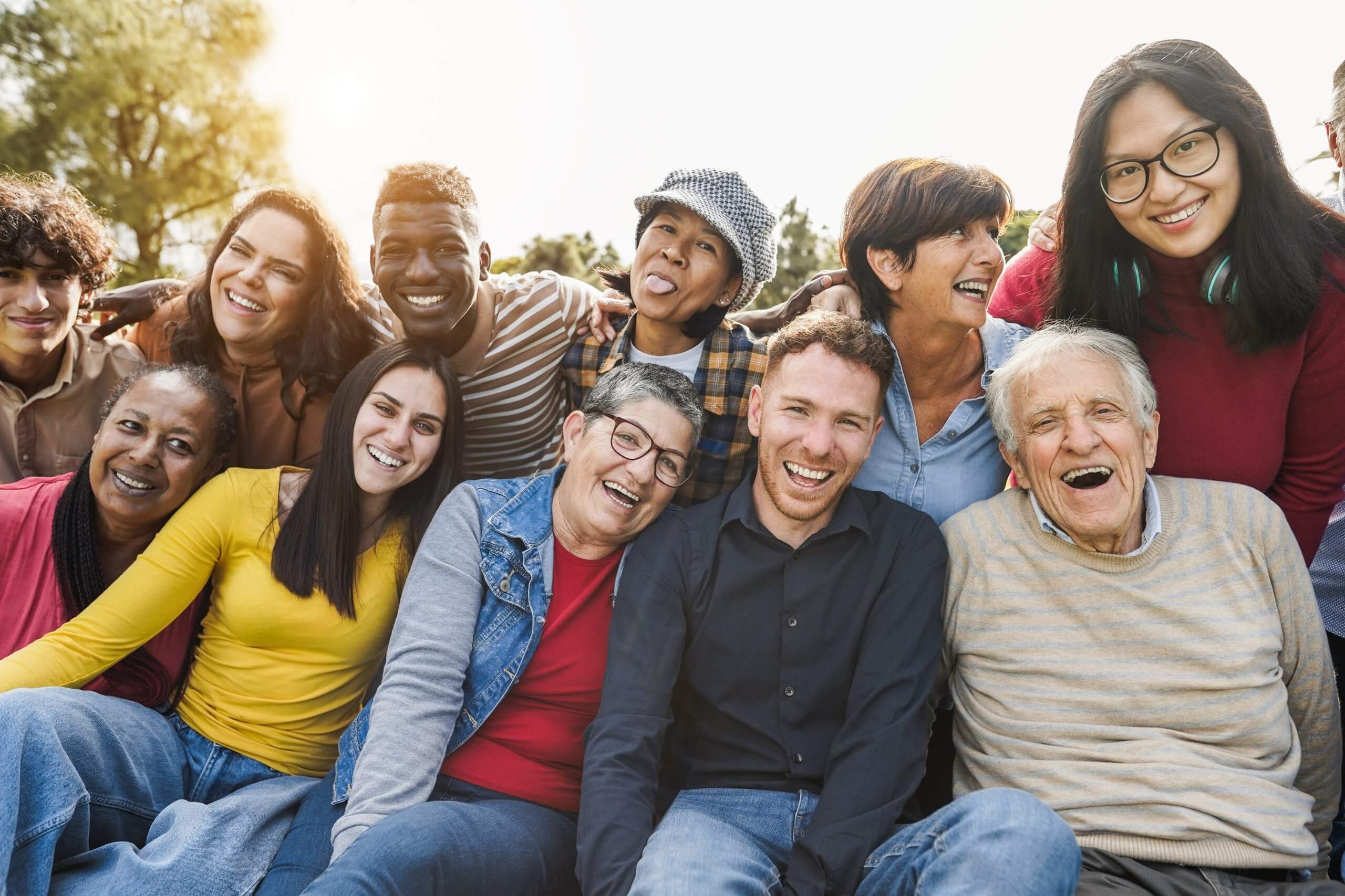 A diverse group of talented people of different ages and backgrounds sit closely together outdoors, smiling and laughing at the camera on a sunny day.