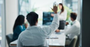 A person seated at a conference table raises their hand, engaging with the culture of open discussion as others listen to a standing woman presenting in a bright, modern office where all figures are out of focus.