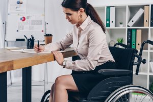 A woman in a wheelchair, dressed in business attire, sits at a desk writing in a notebook. She appears focused—a dedicated government employee with office shelves and a whiteboard visible in the background.