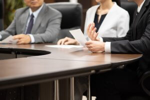 Three people in business attire sit at a conference table during a public sector meeting. One person holds papers and gestures while speaking. The focus is on their hands and the table, with faces not fully visible.