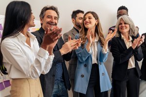 A diverse group of professionally dressed people stand indoors, smiling and clapping as if celebrating or applauding a government achievement.