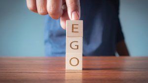 A hand arranges three wooden blocks stacked vertically on a table, each block displaying a letter to spell the word "ego." The background is blurred, emphasizing the concept of ego as the finger lightly touches the top block.