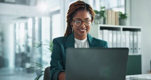 A woman wearing glasses and a green blazer smiles while working on a laptop in a modern public sector office with shelves, binders, and plants in the background.