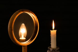 A lit candle stands beside a round mirror. The mirror reflects the candle’s flame, but the reflection appears as an electric light bulb—hinting at how ego can transform a simple reality into something grander. The dark background highlights this illusion.