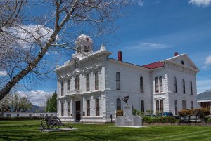 A historic white courthouse with a red roof stands on a green lawn under a blue Orlando sky. A tree with bare branches, a cannon, and a soldier statue monument are in the foreground.