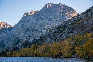A lake bordered by autumn-colored trees sits at the base of a rocky mountain under a clear blue sky—far from Orlando, with sunlight highlighting the mountain’s rugged texture.