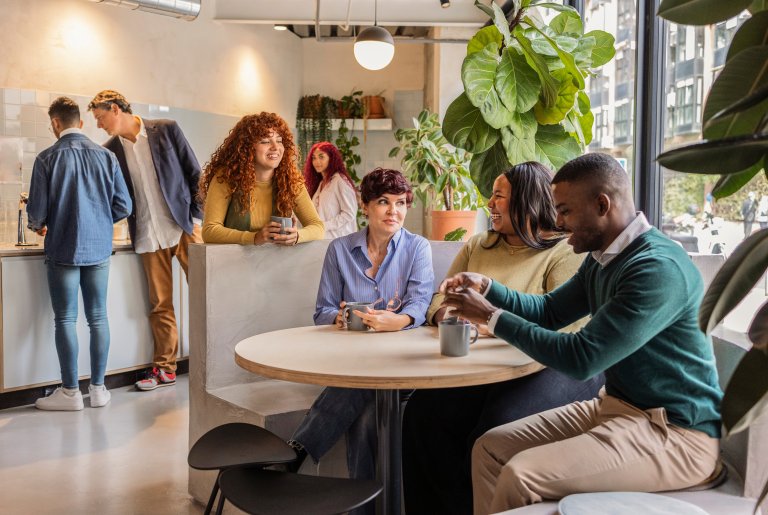 A group of people sit and stand in a modern, bright café. Three people chat at a round table near a window, while others talk and prepare drinks at the counter. Lush plants and vibrant café culture decorate the inviting space.