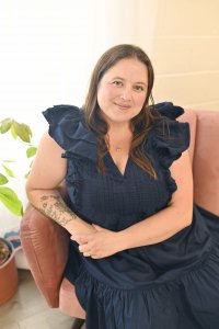 A woman with long brown hair, wearing a navy blue ruffled dress, sits on a pink chair. She has a tattoo on her left forearm and smiles gently, with a potted plant visible nearby.