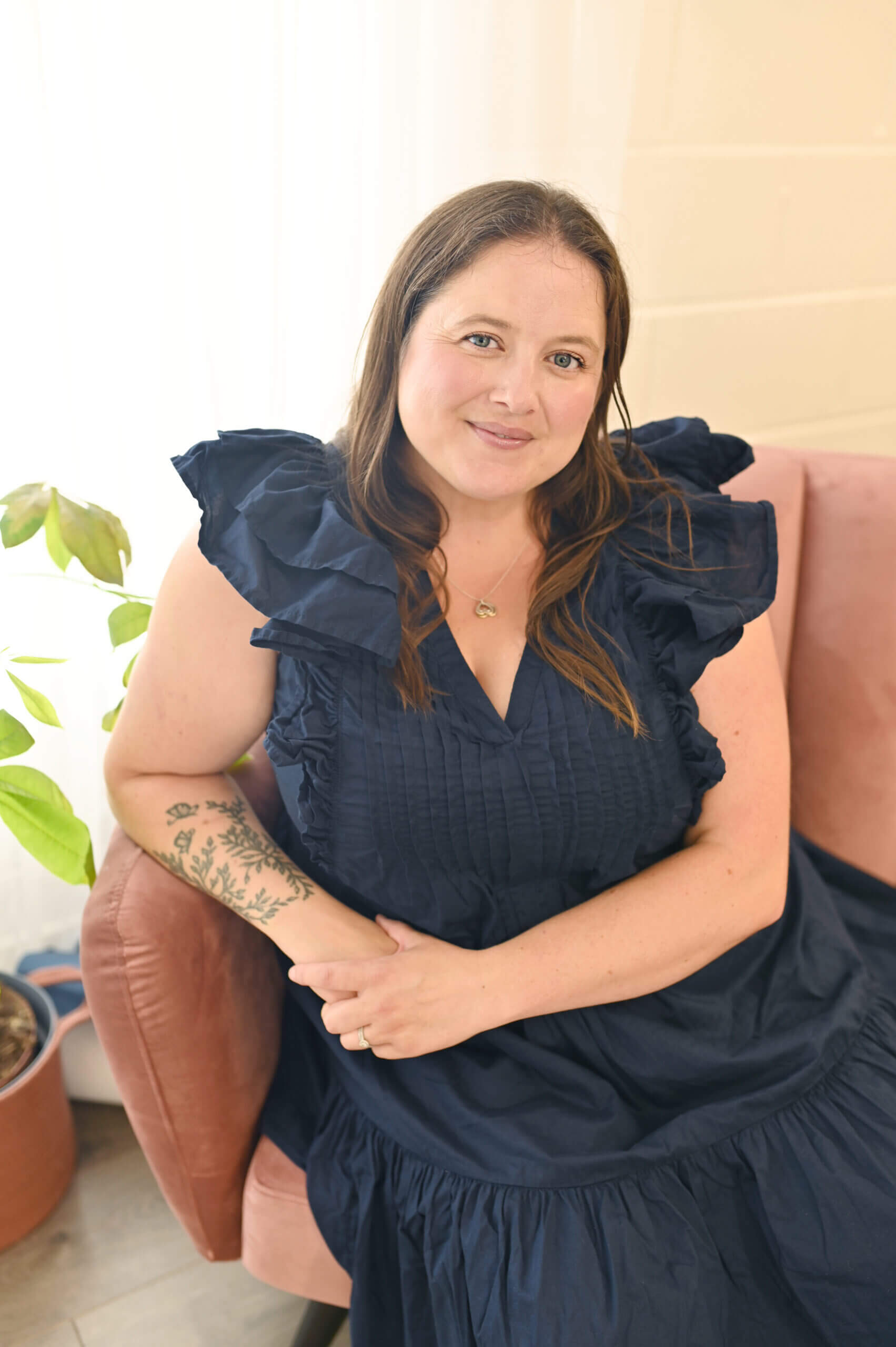 A woman with long brown hair, wearing a navy blue ruffled dress, sits on a pink chair. She has a tattoo on her left forearm and smiles gently, with a potted plant visible nearby.