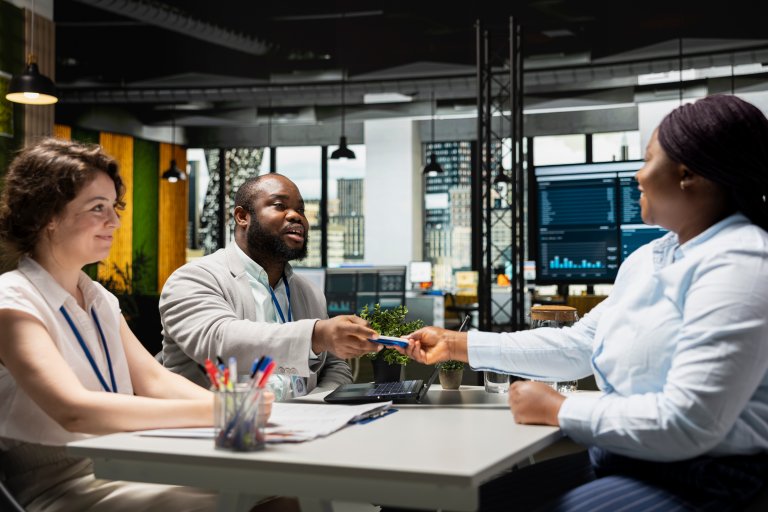 Three government employees sit at a desk in a modern office. A man in a suit hands a document to a woman across the table, while another woman smiles and observes. Laptops, papers, and plants are on the desk.