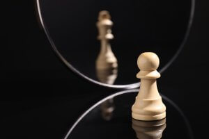 A white chess pawn stands before a round mirror, its reflection appearing as a white king—symbolizing aspiration, feedback, and self-perception—on a glossy black surface with a dark background.