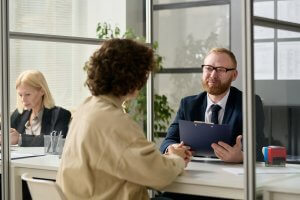 A man in a suit and glasses interviews a person with curly hair across a desk, holding a clipboard—illustrating a government recruitment process. Another woman works at a nearby desk in the modern office setting.
