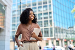 A smiling woman with curly hair stands outside an office building, holding a tablet and looking to the side with quiet ego. She wears a brown shirt and beige pants, with a cityscape reflected in the glass windows behind her.