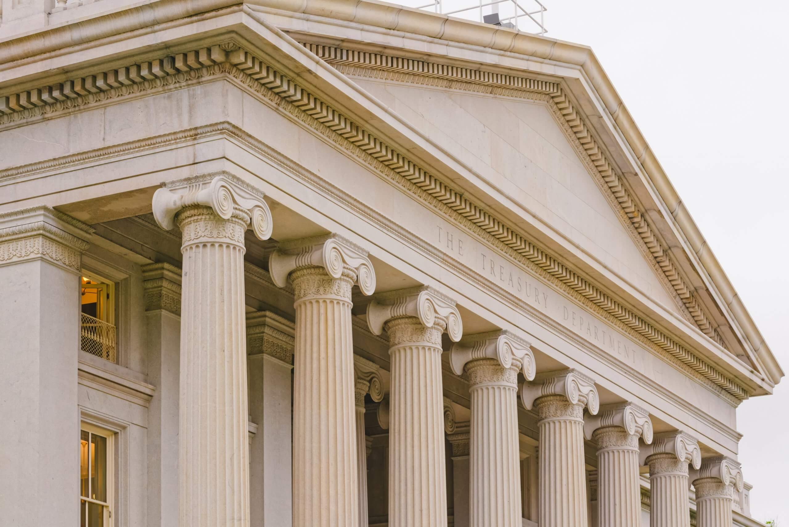 The image shows the neoclassical facade of a federal government building with large columns and the words "The Treasury Department" engraved above them.