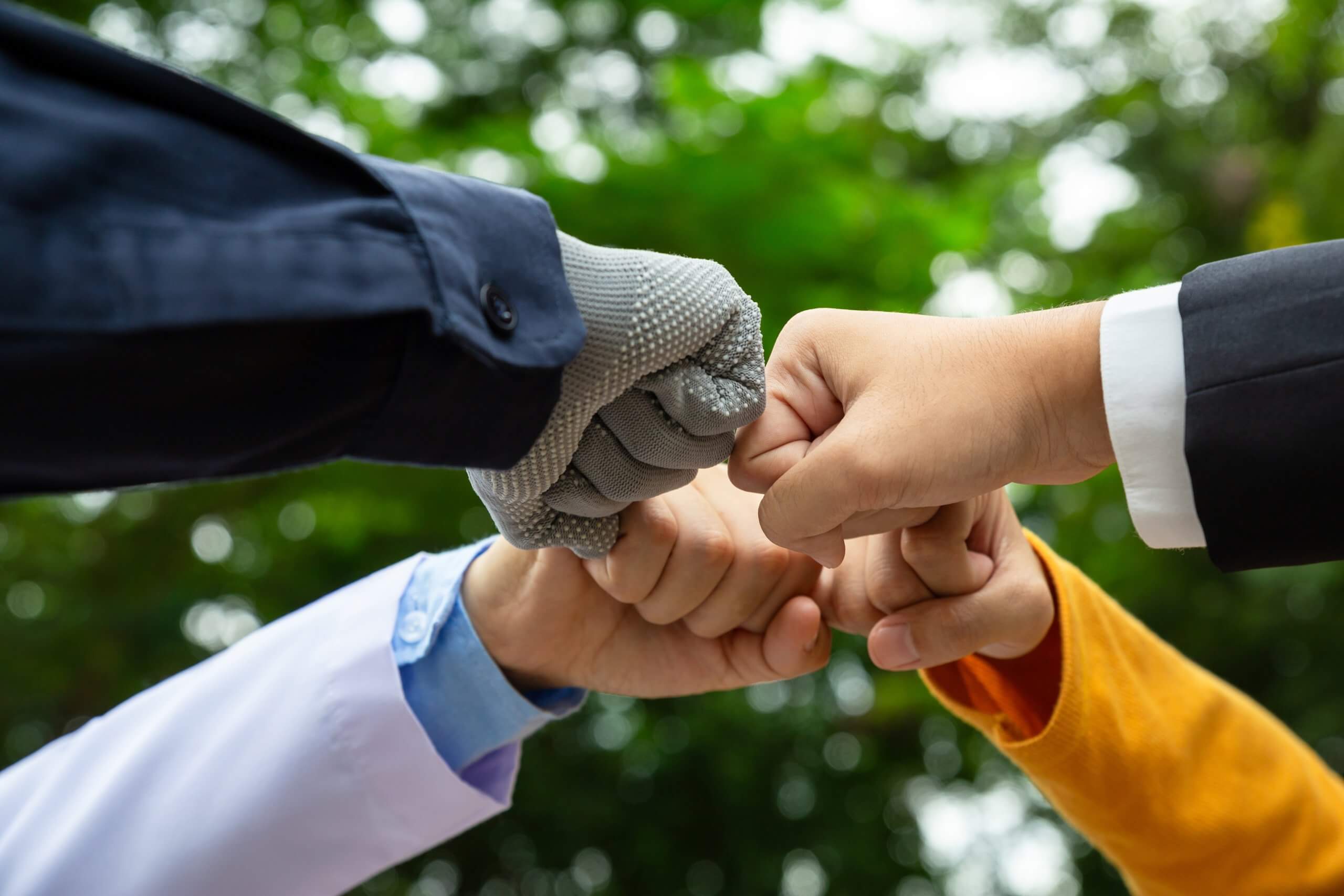 Four hands, each belonging to different generations and wearing variously colored sleeves—one with a gray work glove—join in a group fist bump outdoors, with blurred green trees in the background.