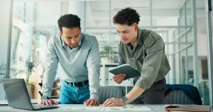 Two employees in business casual attire stand at a desk in a modern office, looking at papers and charts spread out before them. One holds a tablet while the other leans forward, both appearing focused and engaged.
