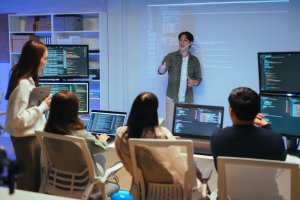 An employee stands and presents code on a projected screen to four seated colleagues, who are looking at laptops displaying programming code in a modern office setting.