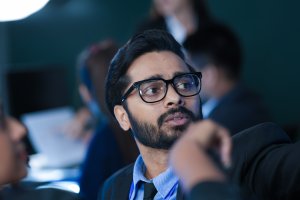 A man wearing glasses and a suit looks to the side while listening during a meeting in an office setting, reflecting on workplace culture as other people are blurred in the background.
