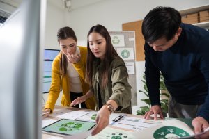 Three people are standing around a desk, reviewing green-colored charts and documents. They appear focused and engaged in discussion, each setting aside ego to effectively collaborate on a project in a modern office setting.
