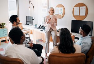 A woman in business attire speaks to four employees sitting in a semi-circle in a modern office, with computers, charts, and documents visible in the background.