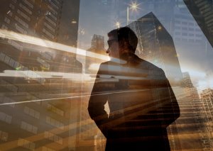 A silhouette of a person in a suit and glasses stands against city skyscrapers with bright light trails, embodying leadership in a dynamic, double-exposure urban scene at dusk.