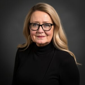 A middle-aged woman with long blonde hair, wearing black glasses, a black turtleneck, and gold hoop earrings, poses against a dark gray background and smiles softly at the camera.
