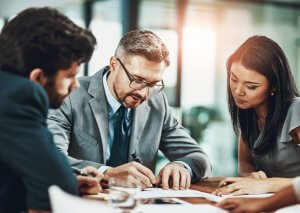 Three business professionals sit at a table in an office, collaborating and reviewing government recruitment documents together. One man is writing while the others pay close attention, all focused and engaged in the discussion.