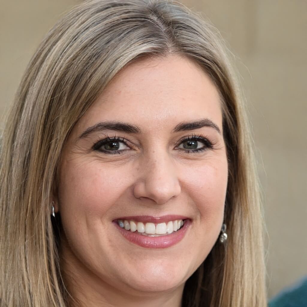 A woman with straight, blonde hair and light skin smiles at the camera. She is wearing earrings and a light-colored top, with a blurred beige background behind her.