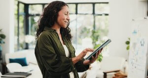 An employee with wavy hair, wearing a green shirt, stands indoors and smiles while using a tablet. She is in a bright room with large windows and plants in the background.