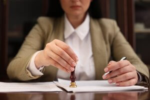 A woman in a blazer presses a wax seal stamp onto a document while holding a pen, honoring traditions passed down through generations as she sits at her desk.