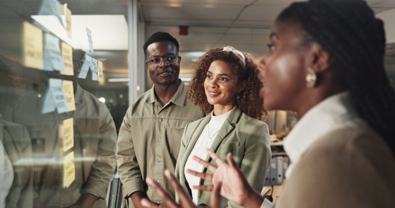 Three employees stand by a glass wall covered with sticky notes, engaged in discussion. One woman speaks while the other two, a man and a woman, listen attentively and smile. The setting appears to be an office.