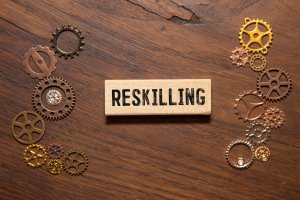 A wooden block with the word "RESKILLING" printed on it sits on a wooden surface, surrounded by small metallic gears and cogs, symbolizing government talent development through skill enhancement.