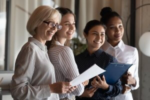 Four women stand together indoors, smiling in a government office setting. One woman holds documents while another has a clipboard, reflecting their professional roles.
