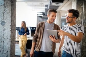 Two men in casual clothes smile and look at a tablet together in a modern office space, highlighting collaboration, while a woman in the background stands near desks and computers.