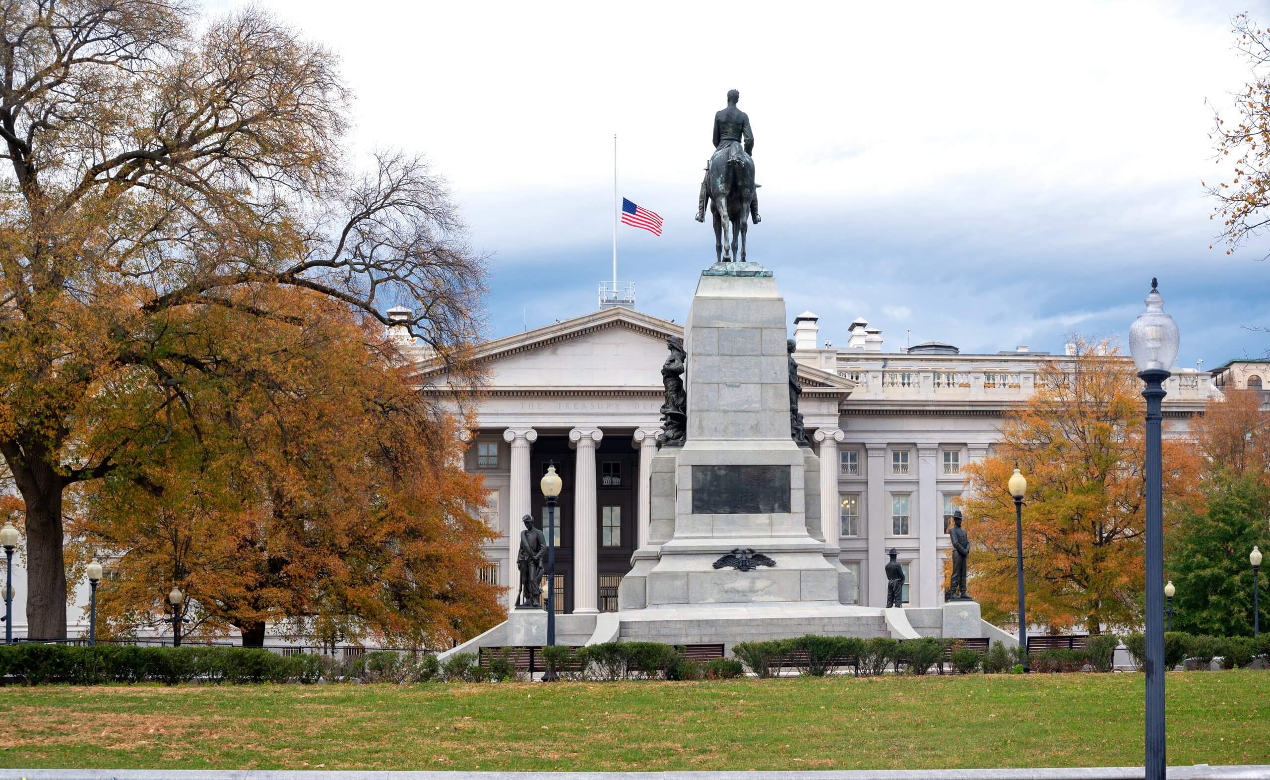 A statue of a person on horseback stands on a stone pedestal in front of a large, columned building with an American flag at half-mast, honoring its dedicated employee. Trees with autumn leaves and lampposts surround the monument.