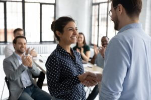 A woman smiles and shakes hands with a man in an office setting, while several colleagues sit at a table in the background clapping and smiling, celebrating her achievement in government leadership.