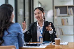 Two women sit across from each other at a table in an office, engaged in lively collaboration. One, wearing a business suit and name badge, gestures enthusiastically as they discuss papers, a tablet, and a coffee cup on the table.