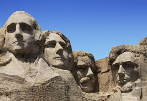 Four large stone sculptures of U.S. presidents' faces, crafted by skilled employee teams, are carved into Mount Rushmore under a clear blue sky. The statues are shown from the shoulders up against the rocky mountain.