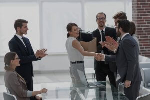 A woman stands smiling and shaking hands with a man while holding a box, surrounded by colleagues applauding in a bright public sector office meeting room.