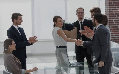 A woman stands smiling and shaking hands with a man while holding a box, surrounded by colleagues applauding in a bright public sector office meeting room.