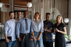 A group of six diverse professionals, some with notebooks and pens, stand and smile in a modern government office, with glass walls and plants visible in the background.