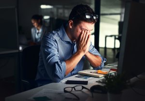 A man in a public service office sits at his desk with his glasses pushed up and his hands covering his face, looking stressed or tired. Papers, a phone, and a computer fill the desk, while another person works in the background.