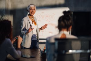 An older woman with short gray hair presents in front of a whiteboard with diagrams, discussing public service strategies with two seated colleagues in a modern office setting.