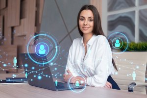 A woman in a white blouse sits at a desk with a laptop, smiling at the camera. Digital network icons and holographic user avatars are superimposed, symbolizing online government communication or connections.