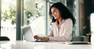 A woman with curly hair, wearing a light pink blouse, is smiling while working on a laptop at a bright, modern desk with large windows—showcasing how leaders thrive in inspiring workspaces.