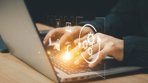Hands typing on a laptop keyboard with glowing digital icons, including gears, padlock, cloud, and AI symbols, representing technology, cybersecurity, automation, and artificial intelligence concepts.