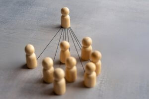 Nine wooden figurines in a group are connected by black lines to a single wooden figurine standing in front, representing leadership or influence within a group—a visual metaphor for public service and guiding others.